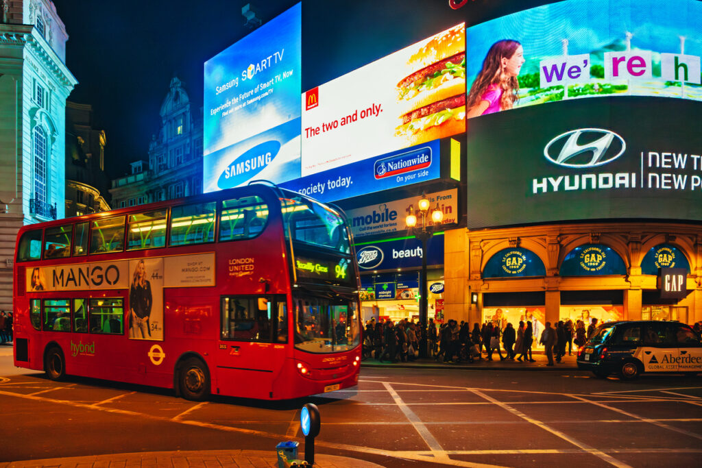 Night view of Piccadilly Circus with illuminated advertising screens, crowd of people, and red double decker bus
